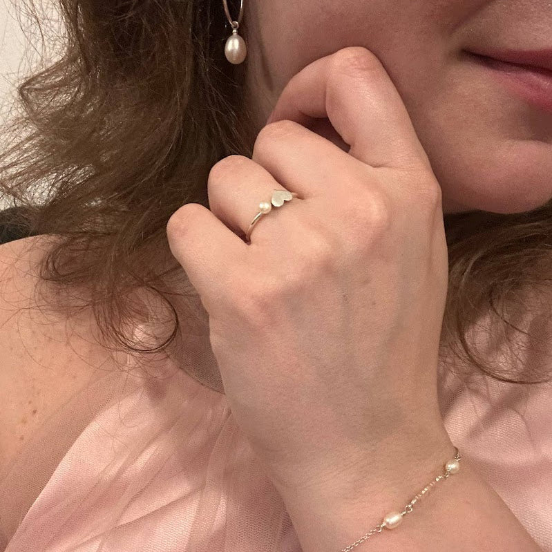 Close-up of a person wearing a pearl earring, ring, and bracelet against a pink background.