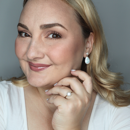 Woman wearing pearl drop earrings and a pearl ring, with a neutral background