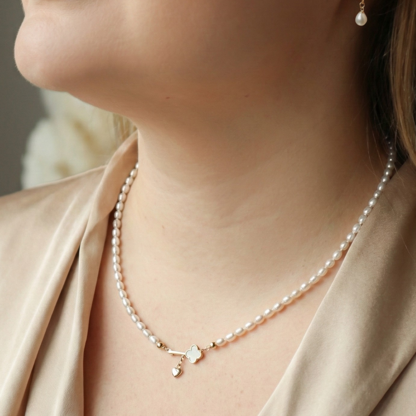 Close-up of a woman wearing a pearl necklace with a key and heart locket charm and pearl earrings with a neutral background